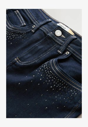 Bootcut denimjeans i mörkblått med en slät textur. Har silverfärgade nitar i ett gradientmönster längs fram.