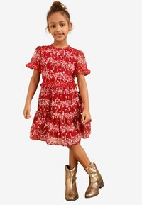 Rotes Kleid mit kurzen, rüschengeschmückten Ärmeln und einem mehrlagigen Rüschenspiegelrock mit pinken Herzmustern. Kombiniert mit schimmernden goldenen Cowboystiefeln.