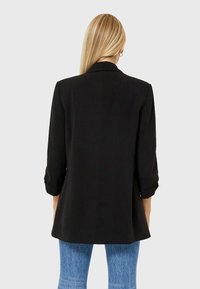 Blazer nero dal design sartoriale con maniche arrotolate. Presenta una texture del tessuto liscia e un retro leggermente più lungo per una silhouette moderna.