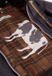 O forro xadrez castanho apresenta uma gráfica de vaca em branco e preto, com um fecho de correr e um detalhe colorido em tom de riscas ao longo da borda.