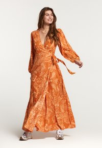 Lange oranje wikkeljurk met ingewikkelde witte bloempatronen, diepe V-hals, lange puffmouwen en een strik in de taille. Geaccentueerd met sneakers.