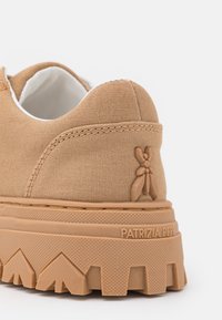 Gros plan sur le talon d'une basket beige avec une semelle texturée, un petit logo abeille, et "PATRIZIA PEPE" embossé sur le bord de la semelle.