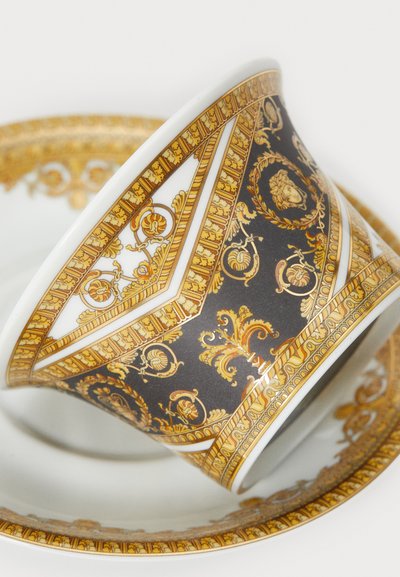 Posliininen teekuppi, jonka muoto on kapeasti kapeneva ja koristeltu yksityiskohtaisilla kullanvärisillä ja tummilla koristekuvioilla. Mukana on samanlainen asetti, jossa on kullanvärisiä yksityiskohtia.