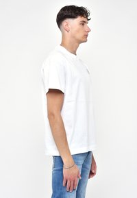T-shirt bianca a maniche corte in cotone, con scollatura rotonda e un piccolo logo sul petto, abbinata a jeans azzurri chiari.
