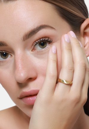 Goldring mit einem wellenförmigen Design, ergänzt durch eine Nahaufnahme einer Hand, die sanft das Gesicht berührt und dabei glatte Haut und neutralen Nagellack zeigt.