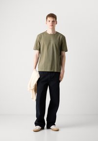 Olivenfarget bomulls-T-skjorte med rund hals, sammen med mørkeblå vide bukser. Modellen holder en lys beige gjenstand i den ene hånden.