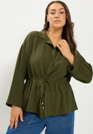Vrouw met lang donker haar die een olijfgroene, gestructureerde blouse met een trekkoord in de taille en blauwe spijkerbroek draagt, poseert voor een effen achtergrond.