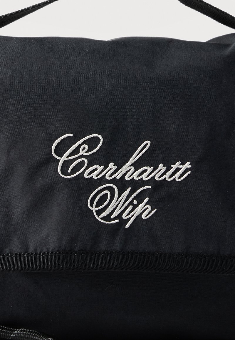 Zwarte stof met witte geborduurde cursieve tekst met de tekst "Carhartt Wip" en een zwarte band zichtbaar aan de bovenkant.