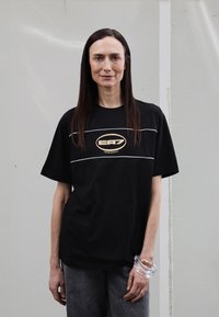 Černé bavlněné tričko s kr átkými rukávy, s horizontálním bílým pruhem a zlatým logem EA7 na hrudi. Nošeno s šedými džíny.