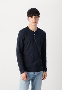Marineblå langermet skjorte laget av myk stoff, med rund halslinje og knappestolpe med hvite knapper. Brukt med lyseblå jeans.