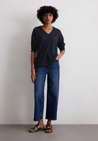 Top nero a maniche lunghe con scollo a V abbinato a jeans a gamba larga blu scuro e sandali neri con fibbia dorata, in piedi contro uno sfondo chiaro.