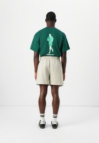 Tricou din bumbac verde cu imprimeu grafic pe spate, asortat cu pantaloni scurți texturați de culoare gri deschis. Pantofi negri și albi cu șosete cu dungi vizibile.