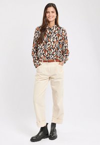 Patterned long-sleeve shirt in black, orange, and cream, paired with cream trousers and black ankle boots, featuring rolled cuffs and a belt.