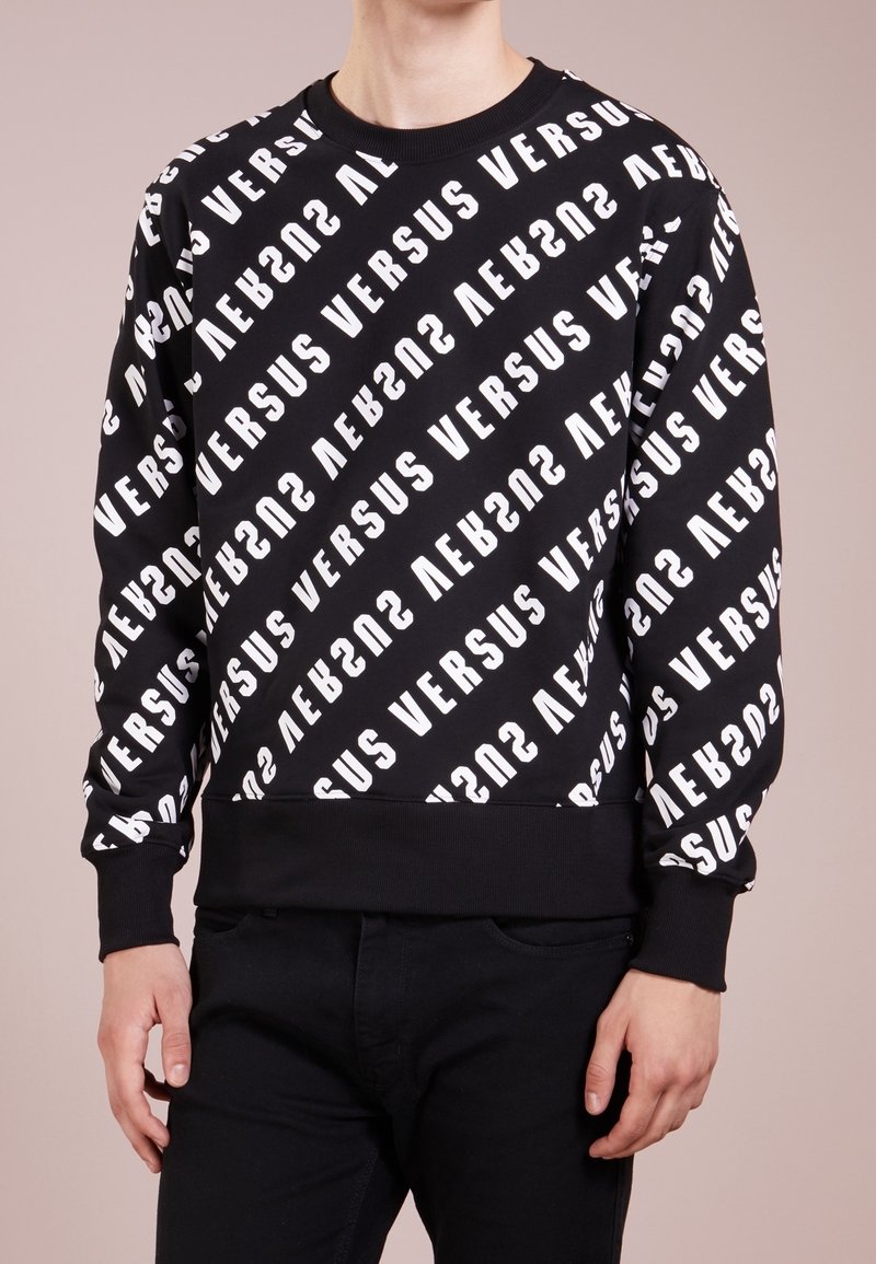 Personne portant un sweat-shirt noir avec un motif de texte diagonal blanc « VERSUS », associé à un pantalon noir, visible du cou aux hanches.