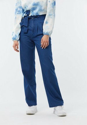 Pantalons en denim bleu à taille haute, avec des plis à l'avant et une coupe large. Associés à un haut en tie-dye bleu clair et des baskets blanches.