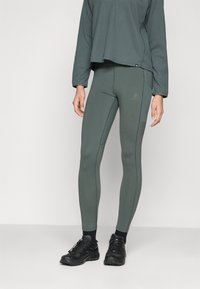 Zöld sportleggingszek, amelyek fitted kialakításúak és kontrasztos varrásokkal rendelkeznek, egy sötétebb zöld árnyalatú, textúrával ellátott, hosszú ujjú toppal párosítva.