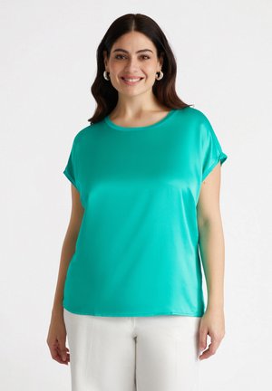Top turchese a maniche corte realizzato in tessuto morbido, con scollatura rotonda e vestibilità rilassata. Indossato con pantaloni chiari.