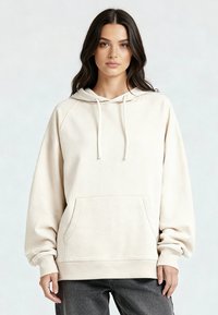 Vrouw met lang donker haar, gekleed in een losse, lichtbeige hoodie met een zak aan de voorkant en trekkoorden, gecombineerd met donkergrijze broek.
