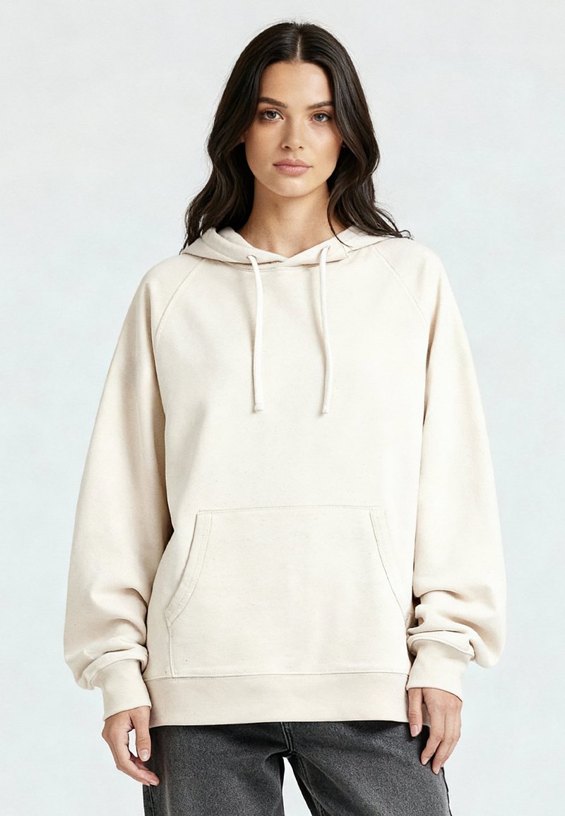 Vrouw met lang donker haar, gekleed in een losse, lichtbeige hoodie met een zak aan de voorkant en trekkoorden, gecombineerd met donkergrijze broek.