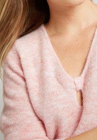 Pull en tricot rose clair avec un col en V, une texture douce et un petit bouton de fermeture au niveau de la poitrine. Les détails incluent des variations de couleur subtiles.