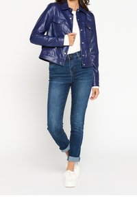 Giacca in finta pelle blu navy con collo, due tasche sul petto e bottoni a pressione argentati, abbinata a jeans blu scuro arrotolati e sneakers bianche.