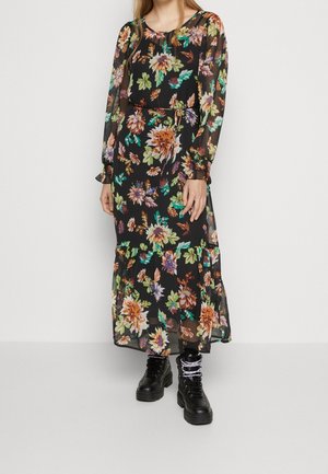 Mujer con vestido largo negro de manga larga con estampado floral, bajo con volantes y botas negras con cordones, de pie contra un fondo liso.