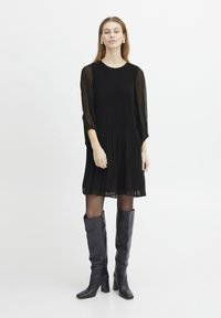 Schwarzes, gefaltetes Kleid mit langen, transparenten Ärmeln, Rundhalsausschnitt und ausgestelltem Saum. Kombiniert mit kniefesten, engen schwarzen Lederstiefeln.
