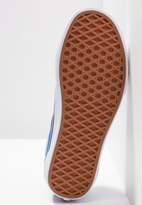 Semelle d'une basket bleue avec des garnitures blanches, montrant une bande de roulement en caoutchouc marron à motif gaufré, positionnée verticalement sur un fond blanc.