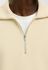 Beige pulovr na zip s texturovaným designem, s vysokým límcem a stříbrným zipem. Nositý přes bílý tričko s kulatým výstřihem.