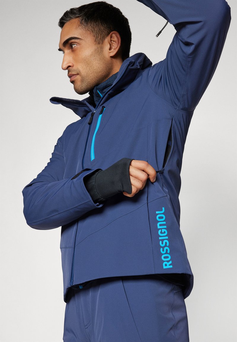 Veste bleu marine avec capuche, dotée d'une fermeture éclair et d'un logo turquoise contrastants. Fabriquée en tissu lisse et résistant aux intempéries avec des poches fonctionnelles.