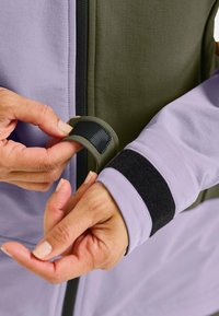 Purple and olive green jacket with a zipper; features Velcro straps on the sleeves. Smooth texture, hands demonstrating closure details.