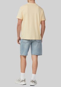 T-shirt en coton jaune clair, coupe décontractée, associé à un short en denim bleu clair. Chaussettes blanches avec des accents gris et chaussures de sport. Fond neutre.