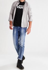 Zwarte T-shirt met witte "VANS" logo, lichtgrijze button-up shirt, blauwe jeans en zwarte gevechtslaarzen. Casual kleding met gelaagde styling.