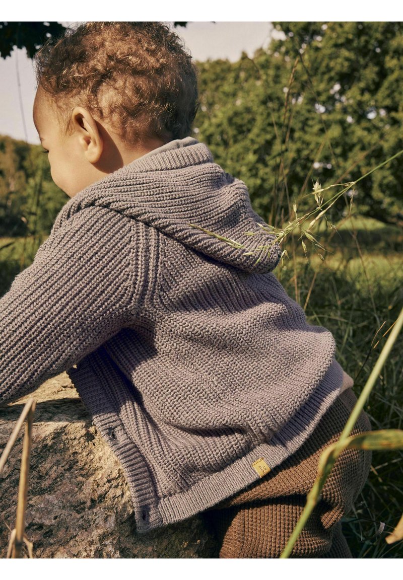 Pull in maglia grigia per bambino con cappuccio, caratterizzato da una lavorazione a costine testurizzata, una piccola etichetta all'orlo e una vestibilità comoda. Pantaloni marroni visibili sotto.