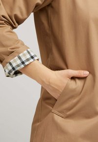 Veste en tissu marron avec un poignet à carreaux retroussé en blanc, vert et noir. Elle dispose d'une poche latérale et d'une texture lisse.