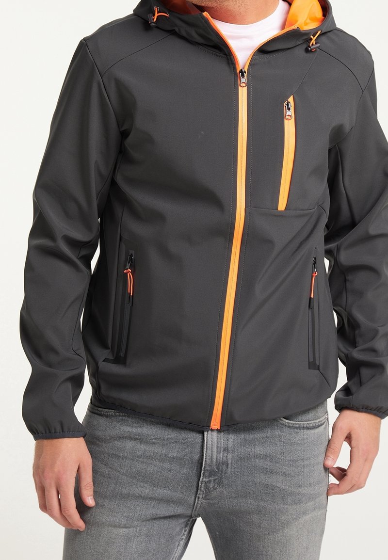 Homme portant une veste noire à capuche avec des fermetures éclair orange et un jean gris, les mains détendues le long du corps.