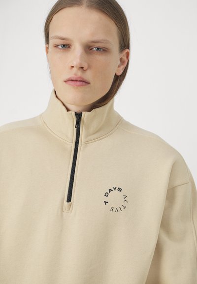 Felpa beige con zip e alto colletto, dotata di logo circolare nero sul petto. Tessuto morbido e testurizzato. Presenta una breve zip nera.
