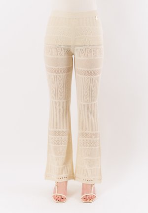 Pantaloni beige a gamba larga lavorati all'uncinetto con motivi intricati e scritte, indossati con tacchi aperti beige chiaro.