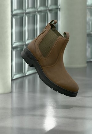 Botte Chelsea en cuir marron avec semelle noire, panneaux latéraux élastiques et languettes, posée devant un mur carrelé brillant et un sol réfléchissant.