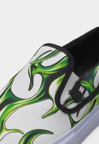 Slip-on αθλητικό παπούτσι με λευκή βάση και πράσινο σχέδιο φλόγας, με μαύρη επένδυση και μια μικρή μαύρη ετικέτα με το λογότυπο DC κοντά στην ελαστική πλευρική ταινία.