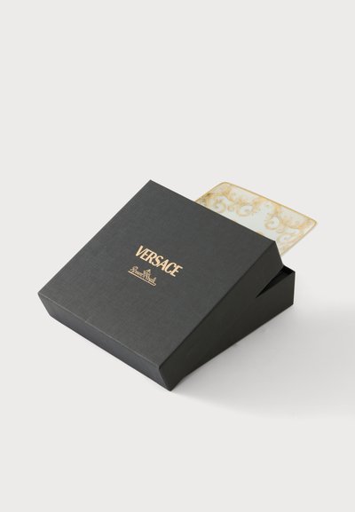 Boîte noire Versace avec logo doré partiellement ouverte, révélant une plaque rectangulaire blanche et dorée ornée à l'intérieur.