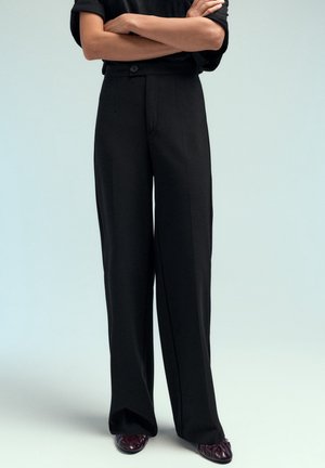 Personne portant un pantalon noir à jambes larges et des chaussures bordeaux, debout les bras croisés devant un fond uni.