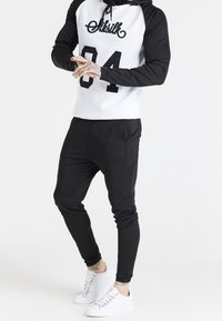 Completo atletico bianco e nero con un top con cappuccio con stampa "Siksilk 84" e pantaloni neri affusolati. Il materiale sembra liscio, con una sottile texture.