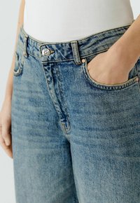 Lichtblauwe denimjeans met vijf zakken, zichtbare stiksels en een zilveren knoopsluiting aan de taille. Glad, licht vervaagd textuur.