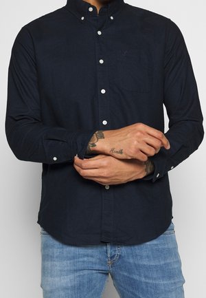 Person trägt ein dunkelblaues Hemd mit Knöpfen und hellblaue Jeans und richtet die Manschette am linken Handgelenk, die ein Tattoo mit dem Wort "Demütig" zeigt.