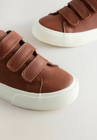 Hnědé kožené boty s třemi suchými zipy a bílými gumovými podrážkami, umístěné na světlém pozadí, které ukazuje jejich designové detaily.