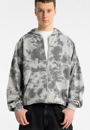 Młody mężczyzna ubrany w szaro-białą bluzę z kapturem na zamek błyskawiczny typu tie-dye z kieszeniami z przodu i czarne spodnie, stojący na jednolitym białym tle.