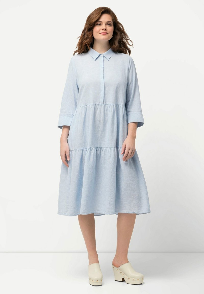 Vestido azul claro con cuello, mangas tres cuartos, frente con botones y falda en capas y fluida. Hecho de una tela ligera.