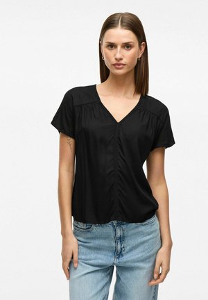 Zwarte korte mouwen top met een V-hals, voorzien van gerimpelde details langs de schouders en een knoopsluiting aan de voorkant, gecombineerd met lichtblauwe jeans.