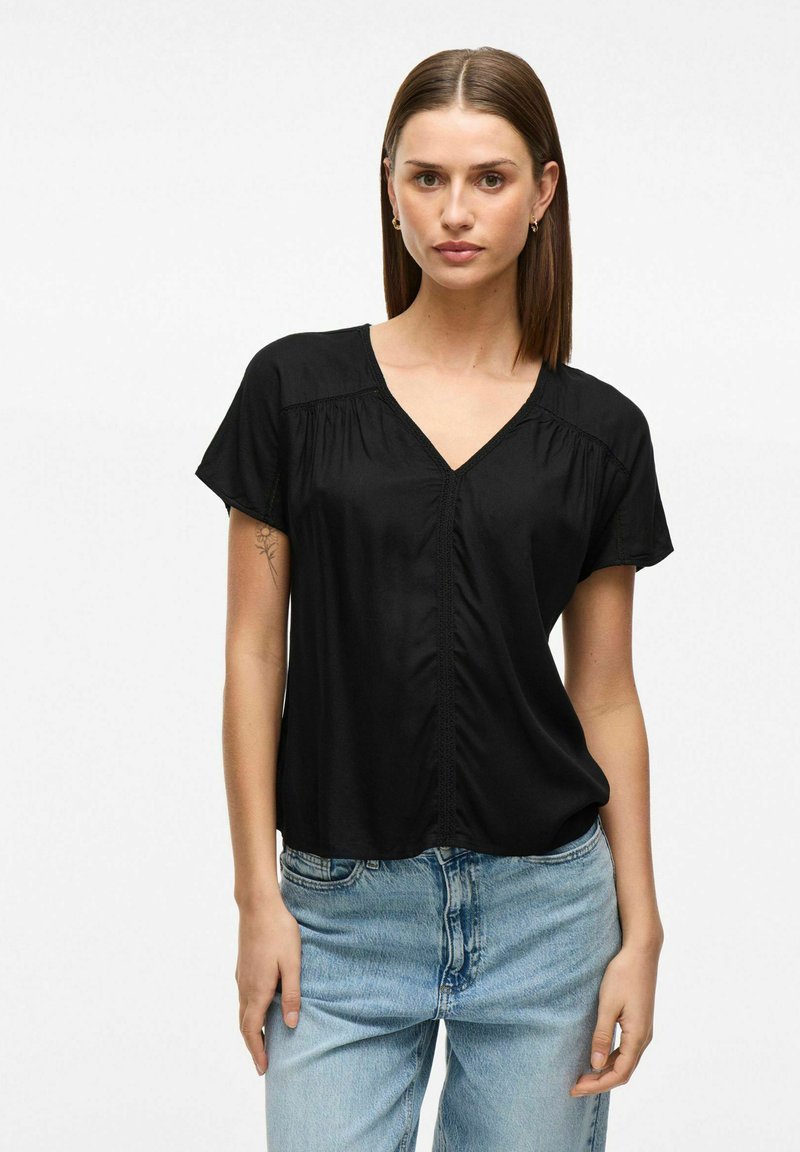 Top nero a maniche corte con scollatura a V, caratterizzato da dettagli arricciati sulle spalle e una patta con bottoni sul davanti, abbinato a jeans azzurri chiari.